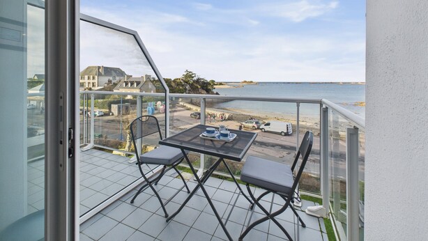 Een balkon met een tafel en stoelen, met uitzicht op een strand en een parkeerplaats.