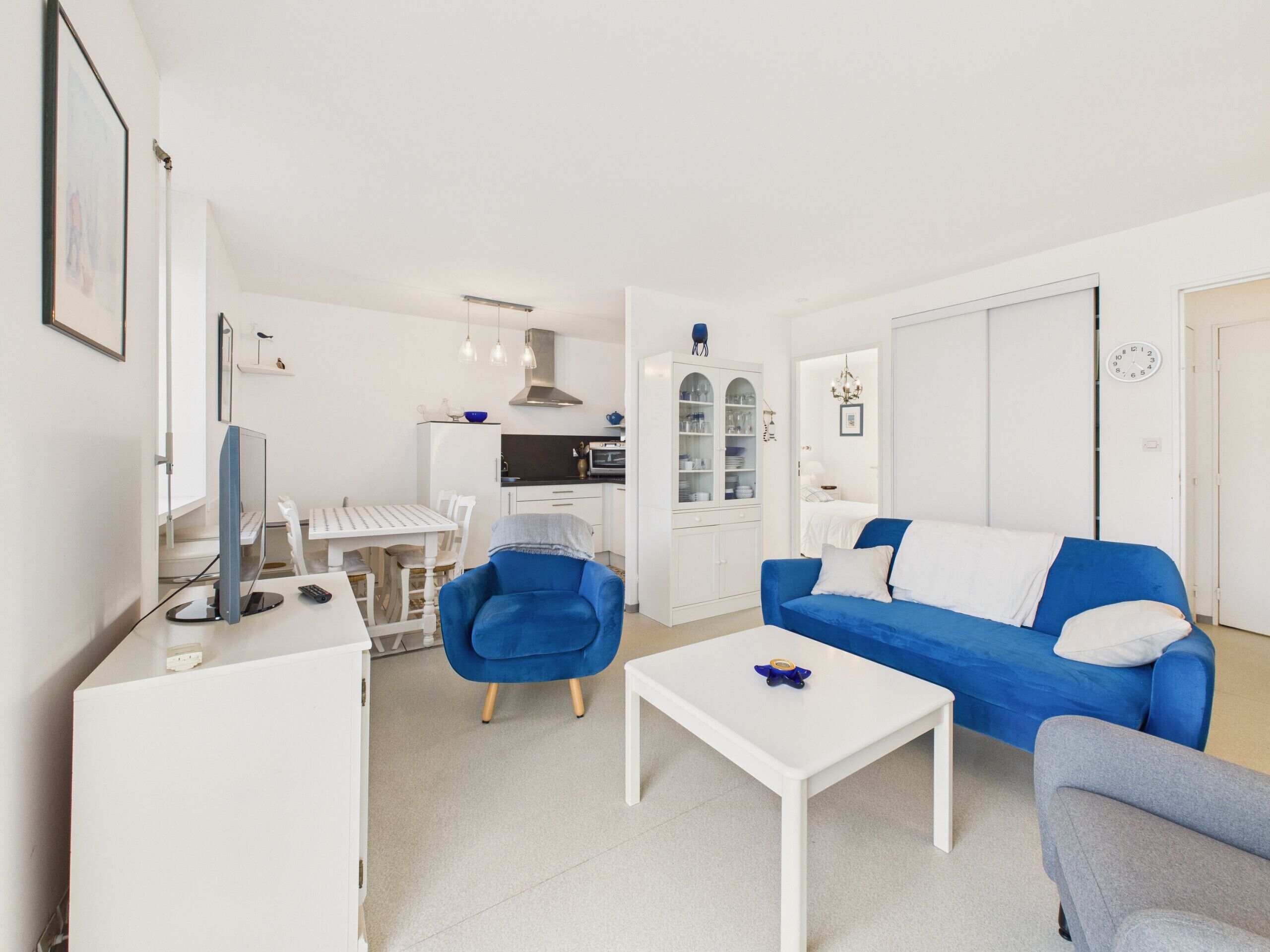 Un salon moderne avec un canapé bleu, une table basse blanche et un fauteuil bleu. On y trouve également un coin cuisine avec des meubles blancs et une table à manger.