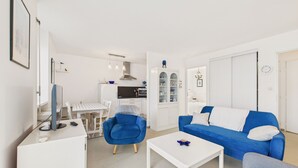 Een moderne woonkamer met een blauwe bank, een witte salontafel en een blauwe fauteuil. Er is een keuken met witte kasten en een eettafel.