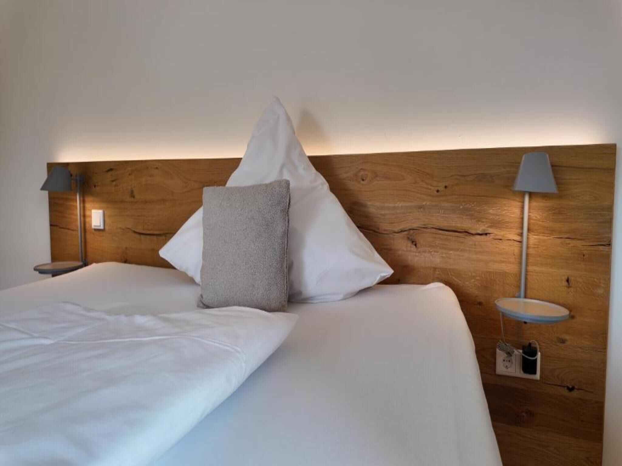 Un lit bien fait, avec du linge de lit blanc et un oreiller gris, flanqué de deux tables de chevet avec des lampes, le tout adossé à une tête de lit en bois.