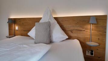 Un lit bien fait, avec du linge de lit blanc et un oreiller gris, flanqué de deux tables de chevet avec des lampes, le tout adossé à une tête de lit en bois.