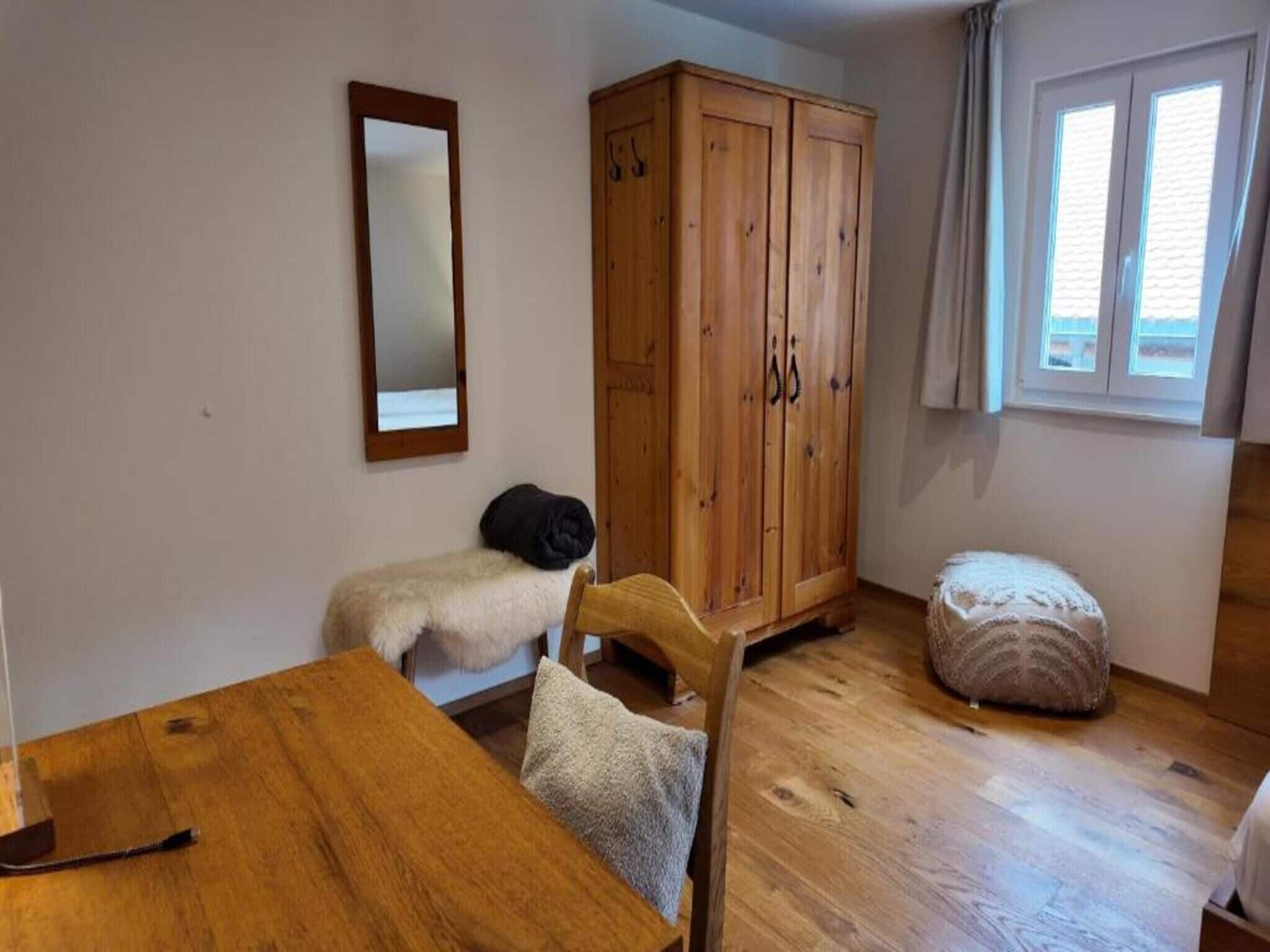 Ein Zimmer mit einem Holztisch, einem Schrank, einem Spiegel und einem Fenster mit Vorhängen.