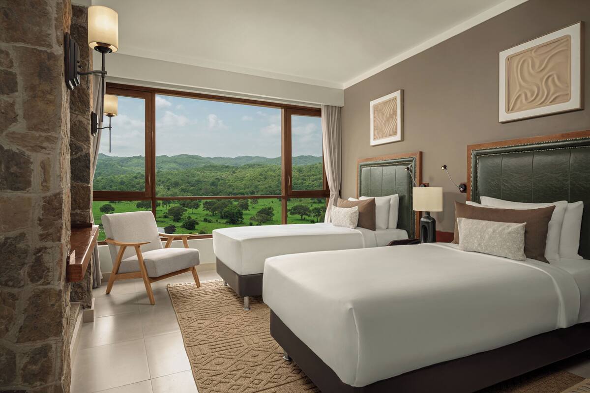 A hotel room with two beds, a large window with a mountain view, a stone wall, and a modern lamp.
