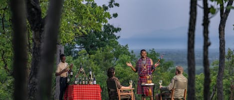 A group of people outdoors with a table set for a meal, surrounded by trees and a view of the landscape.