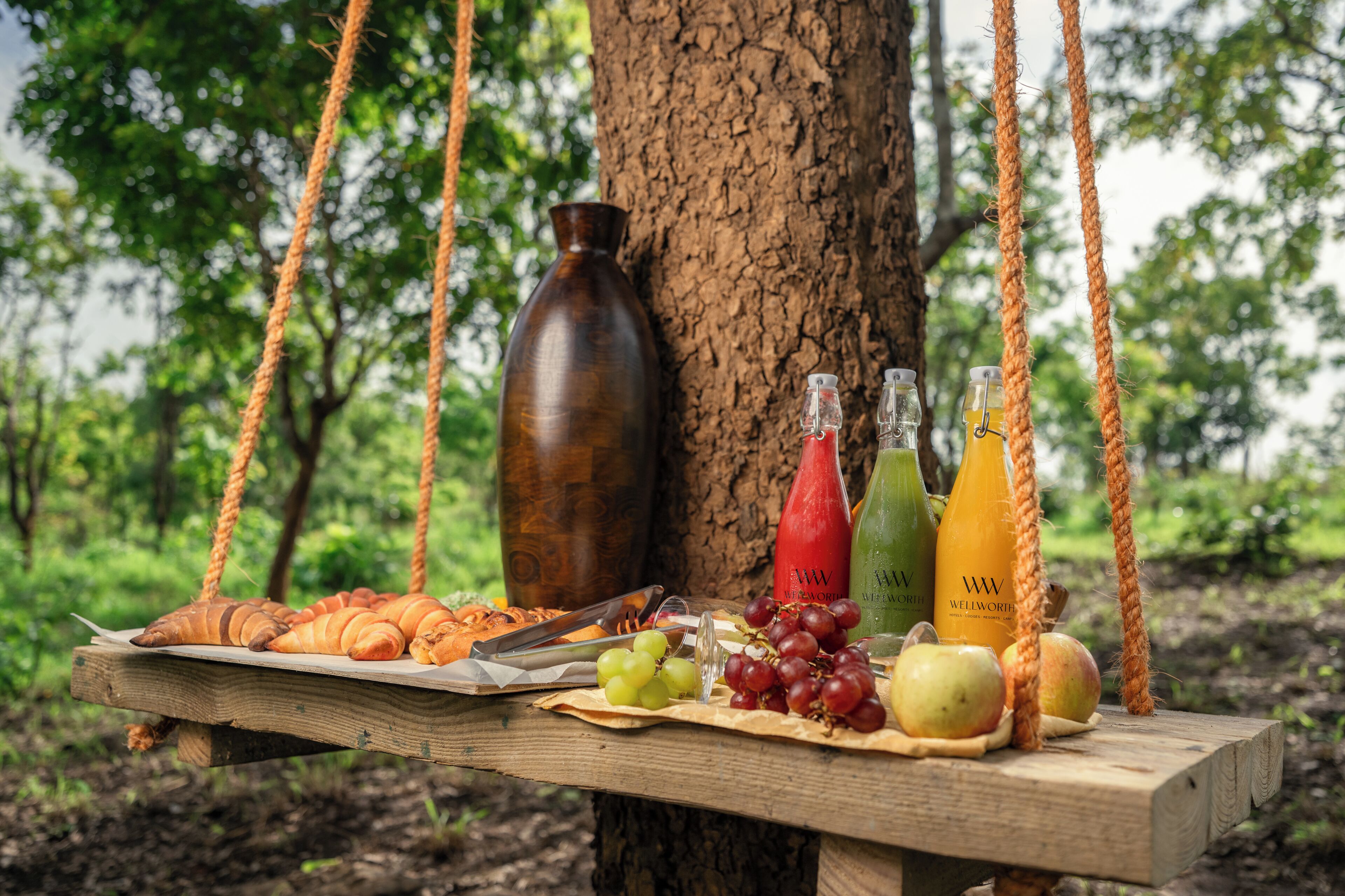 A wooden table with food items and drinks, suspended by ropes, against a tree backdrop.