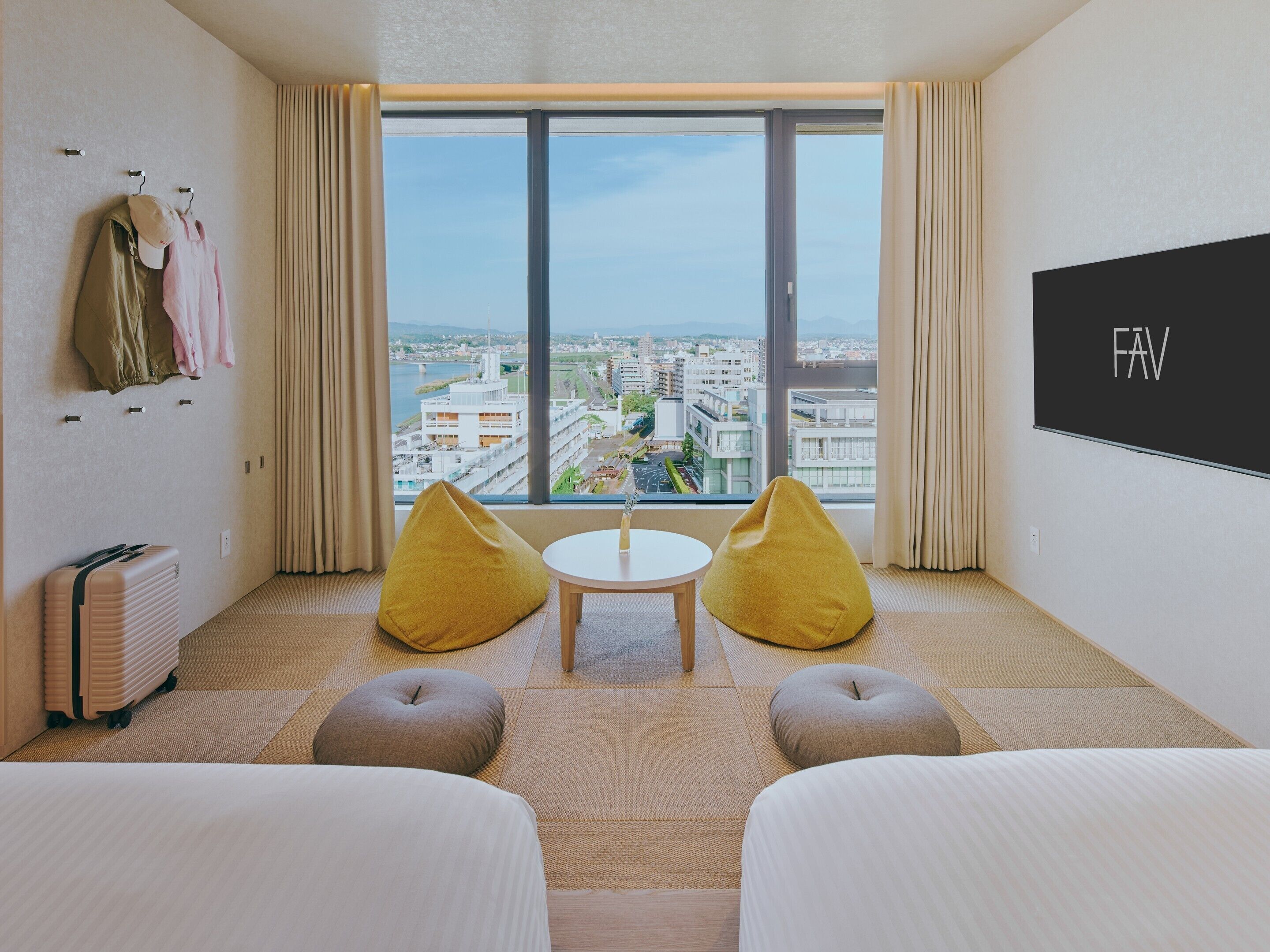 Quarto de hotel com uma janela grande, dois puffs, uma cama, uma mesa e uma televisão na parede.