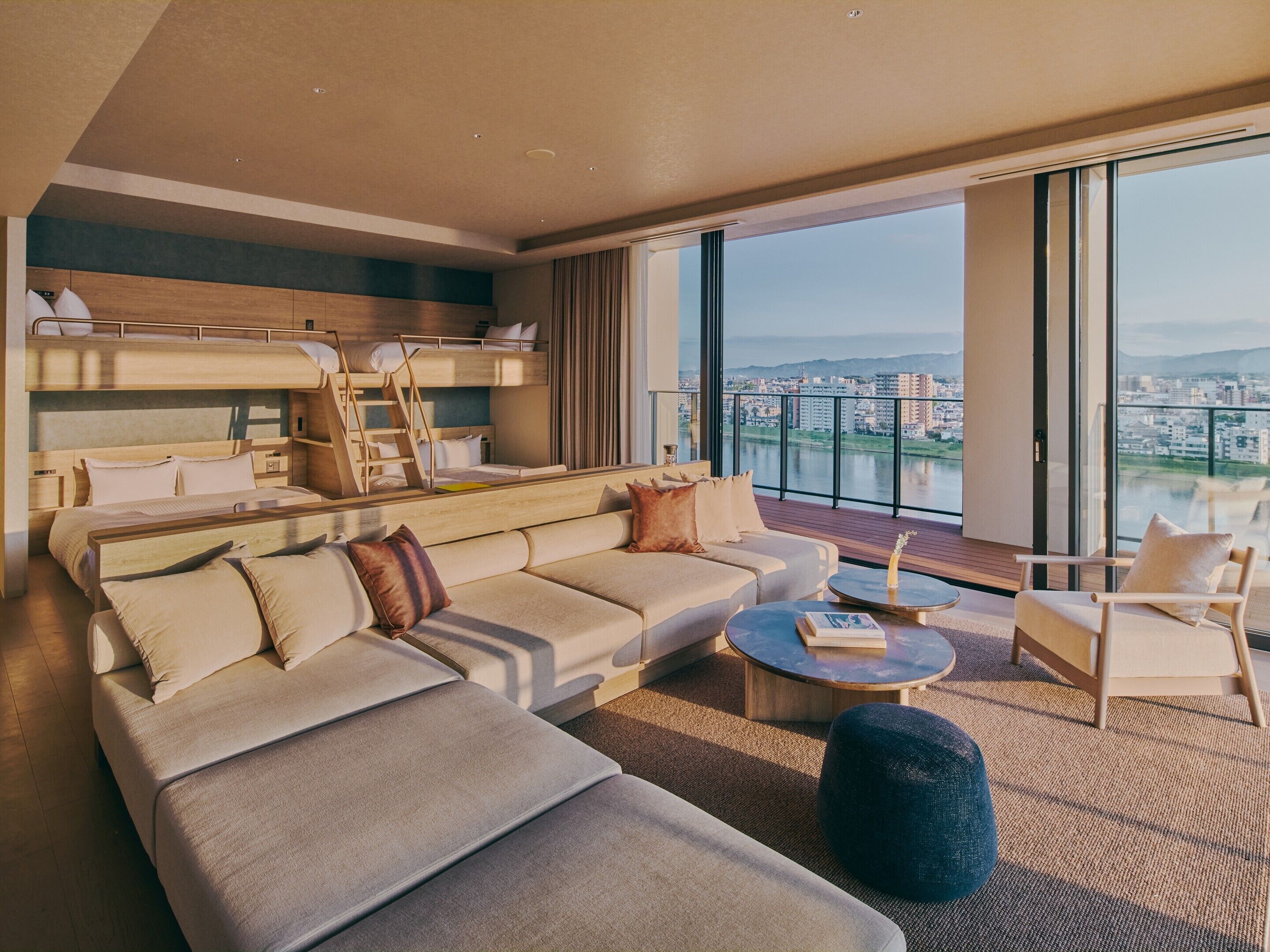 A modern hotel room with a large sofa, a small round table, a bunk bed, and a balcony with a city view.