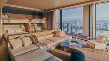 A modern hotel room with a large sofa, a small round table, a bunk bed, and a balcony with a city view.