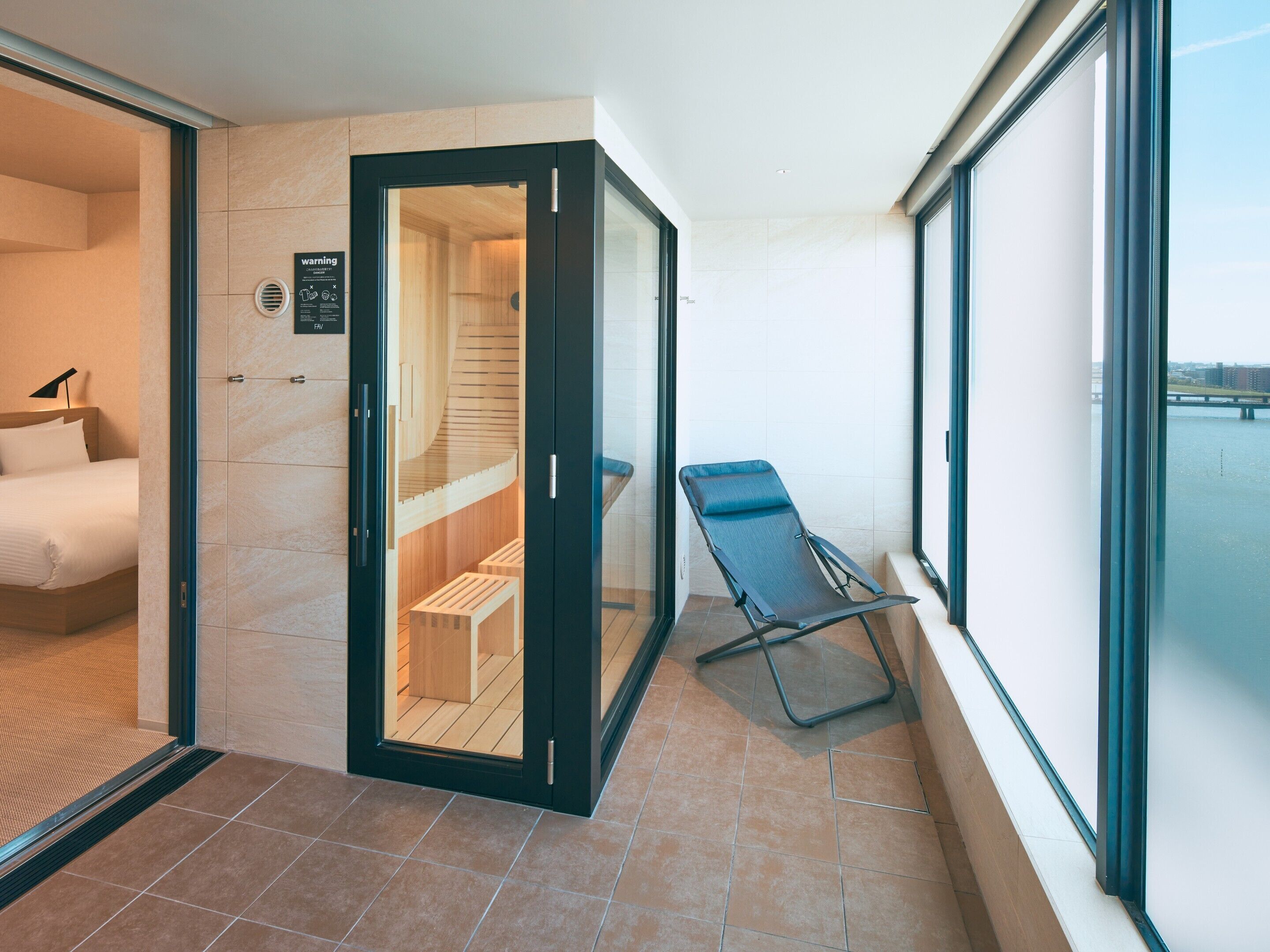 A modern hotel room with a wooden sauna, a bed, and a view of the water through large windows.