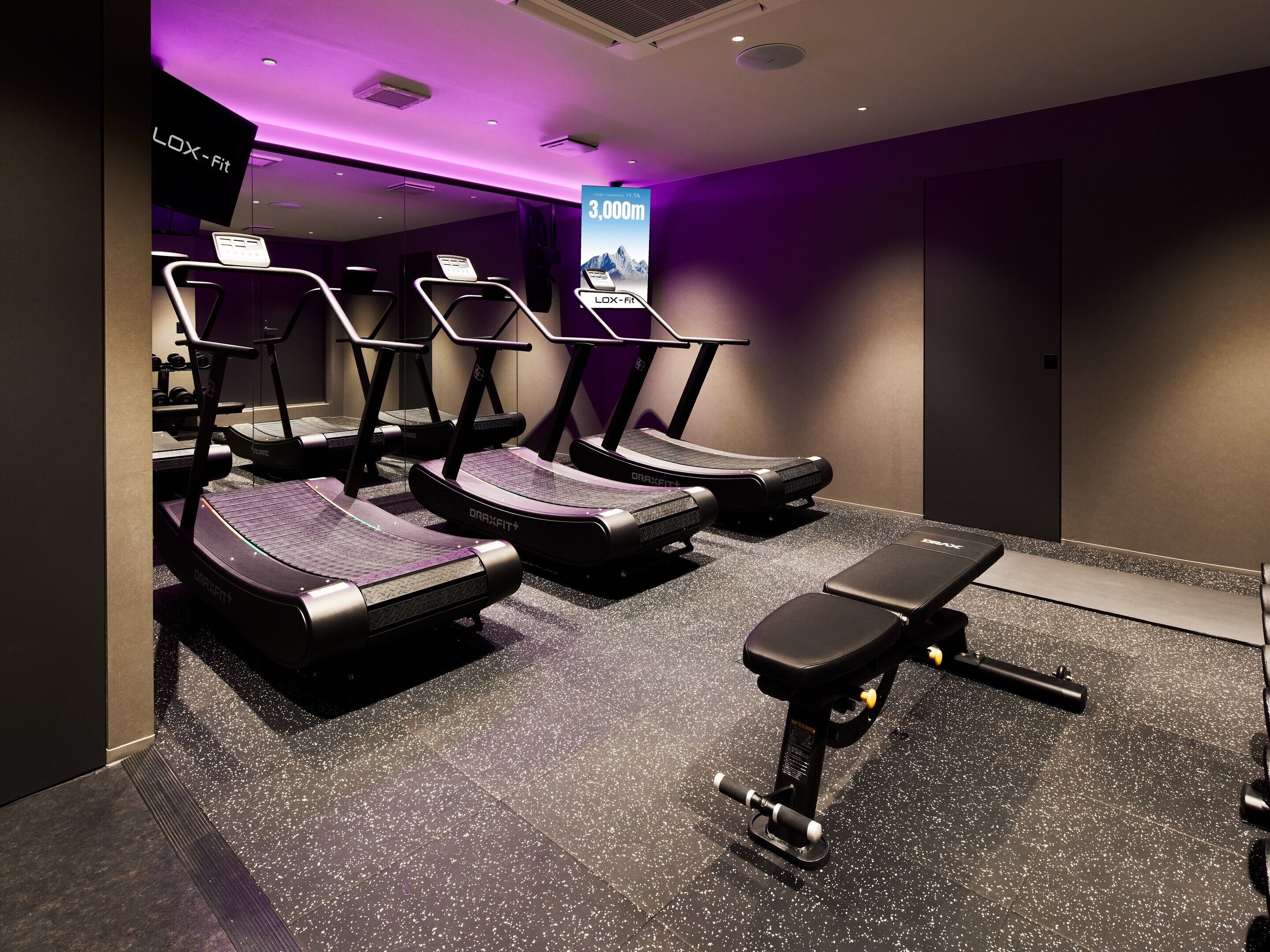 A gym with treadmills, a bench, and a wall-mounted screen displaying '3,000m'.