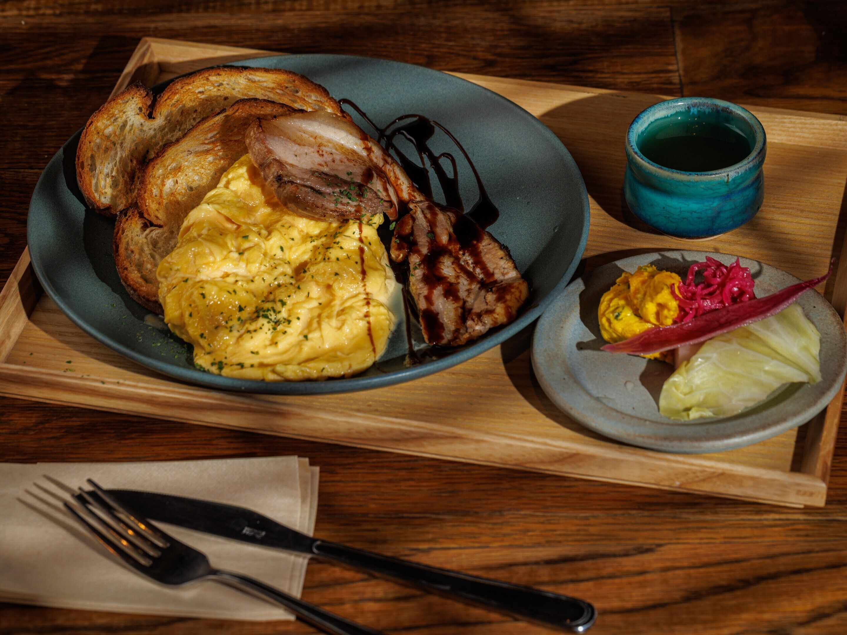 A breakfast plate with toast, scrambled eggs, and bacon, a small plate with pickles and a red pepper, and a blue mug on a wooden tray.