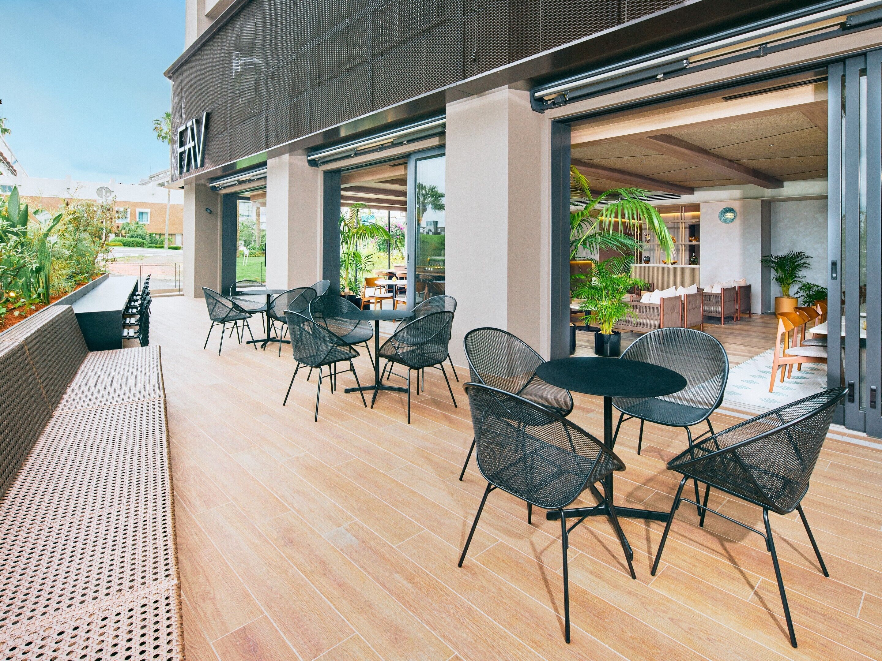 An outdoor patio with seating and tables, a covered area with a view of the garden, and a building entrance.