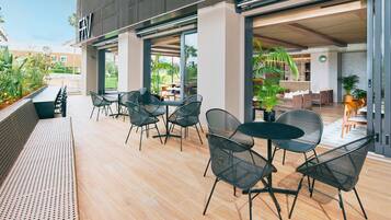 An outdoor patio with seating and tables, a covered area with a view of the garden, and a building entrance.