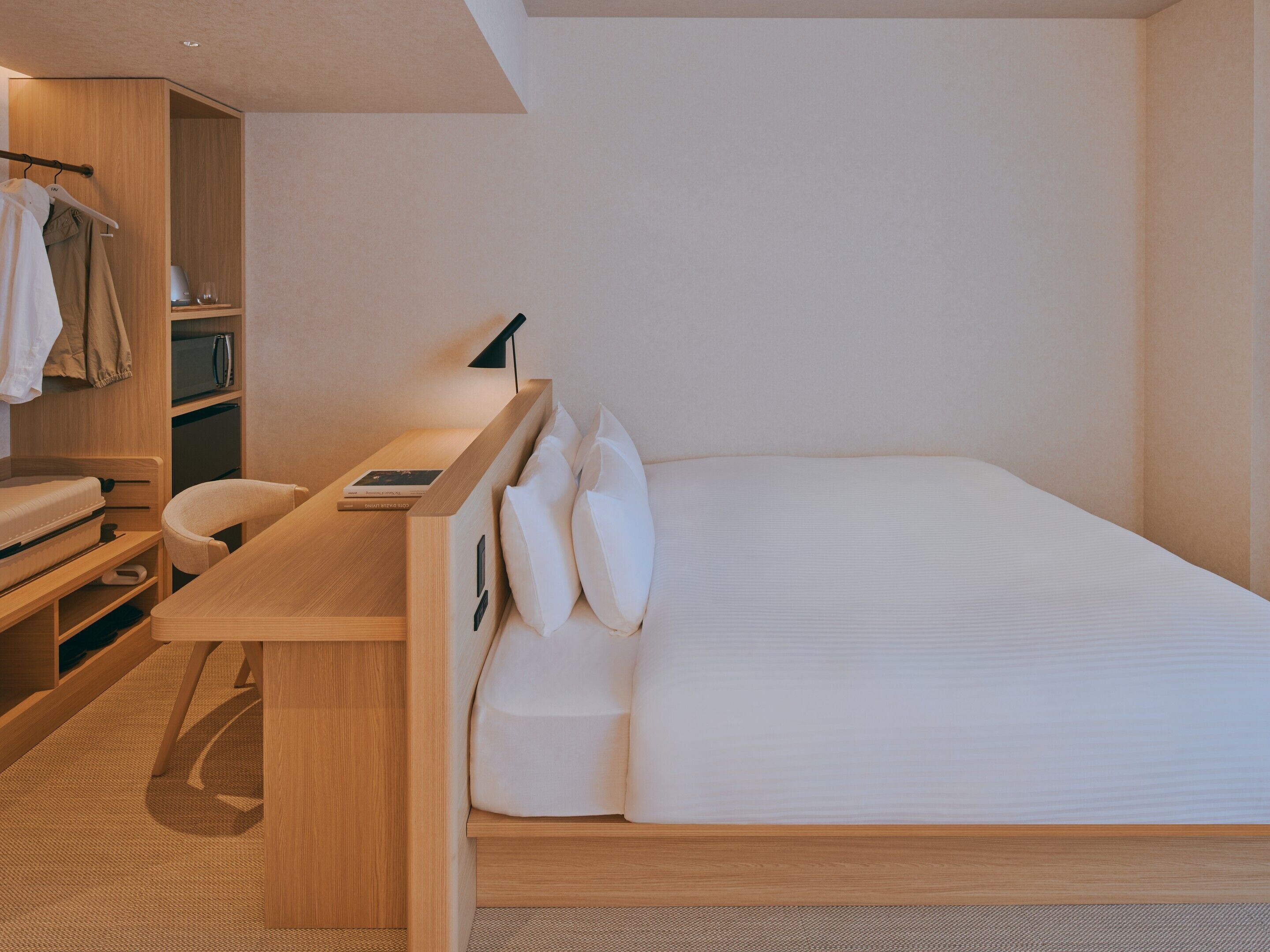 A compact hotel room with a bed, a desk, a chair, a wardrobe, and a bedside table.