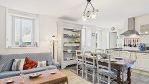 En hyggelig stue med sofa, spisebord og stole, samt et køkken med hvide skabe og en hængende lampe.