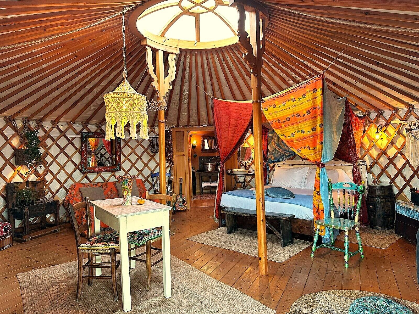 A yurt interior with a central bed, a hanging lamp, a table with chairs, and a small kitchen area.