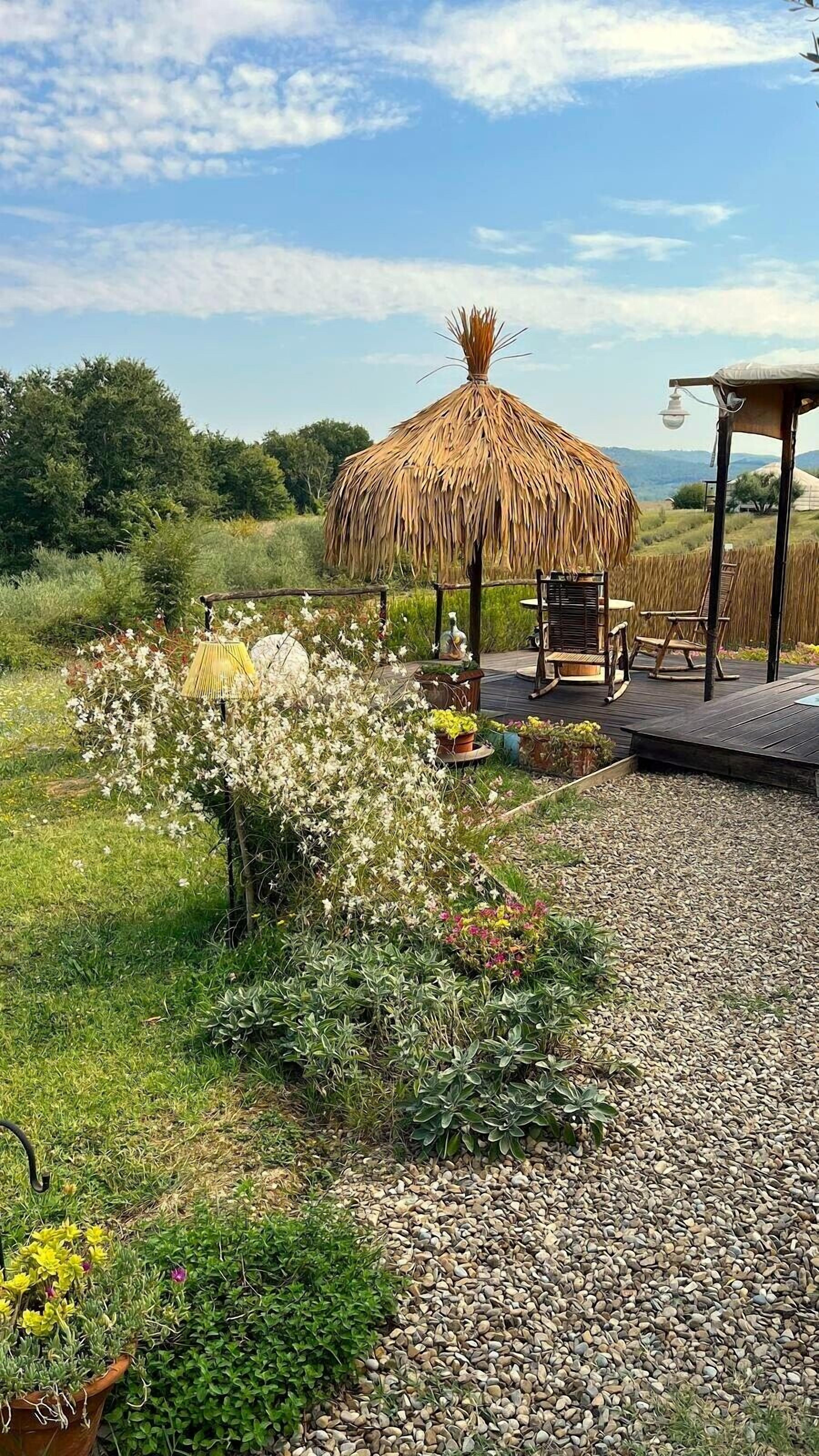 A thatched-roof gazebo with a wooden deck, surrounded by a garden with various plants and flowers.