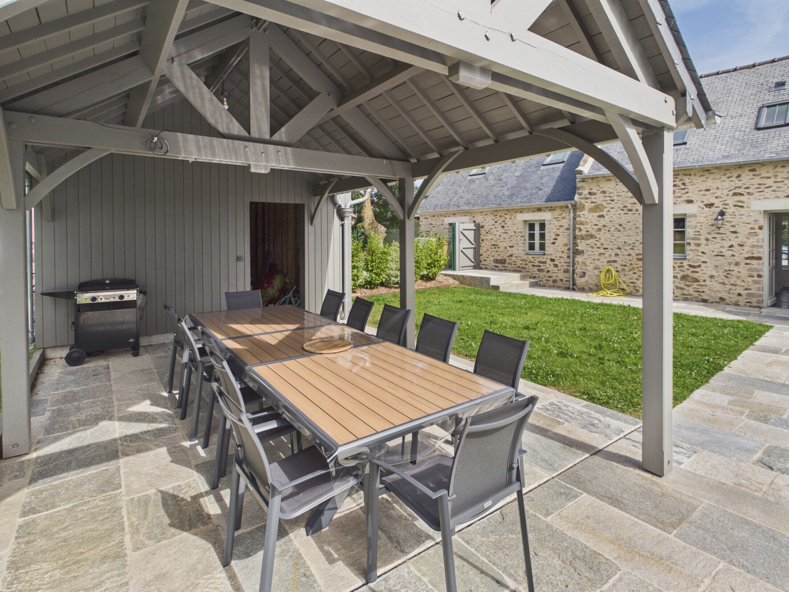 Een overdekt terras buiten met een tafel en stoelen, een barbecue en een stenen gebouw op de achtergrond.