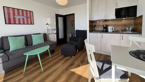 Ein kompakter Wohnbereich mit einer Couch, einem kleinen Esstisch, einer Küchenzeile und einer Tür zu einem weiteren Raum.