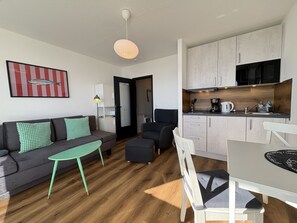 A compact living space with a sofa, a small dining table, a kitchenette, and a door leading to another room. - Strandhotel - 19 "Sardine tin" - More sea views from the "sardine tin"! (Dahme)