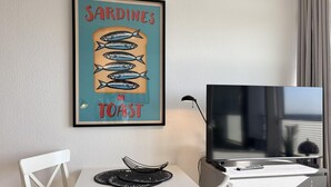 Ein Zimmer mit gerahmtem Poster von Sardinen auf Toast, einem Fernseher, einem Schreibtisch mit Stuhl und einem Fenster mit Vorhängen.