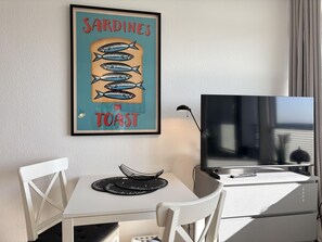 A room with a framed poster of sardines on toast, a TV, a desk with a chair, and a window with curtains. - Strandhotel - 19 "Sardine tin" - More sea views from the "sardine tin"! (Dahme)