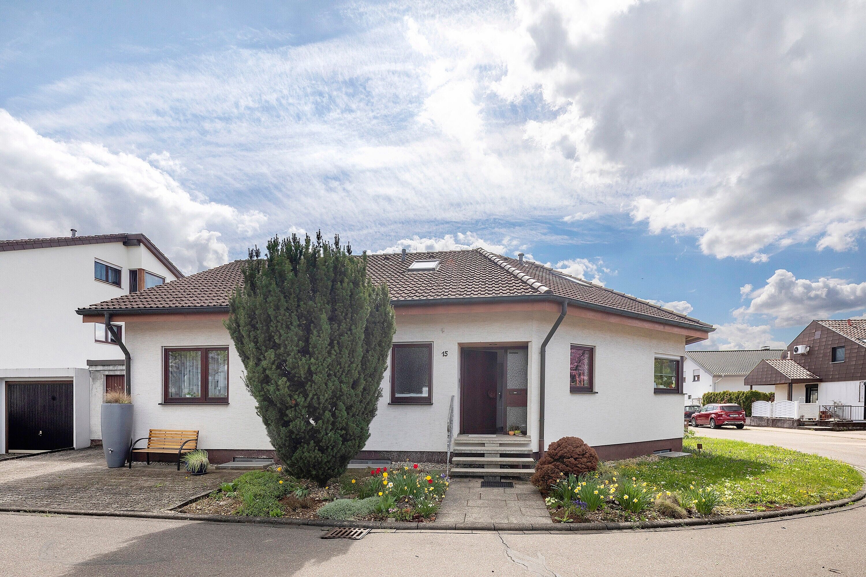 Een gelijkvloers huis met een veranda, een garage en een verzorgde tuin met bloemen en een boom.