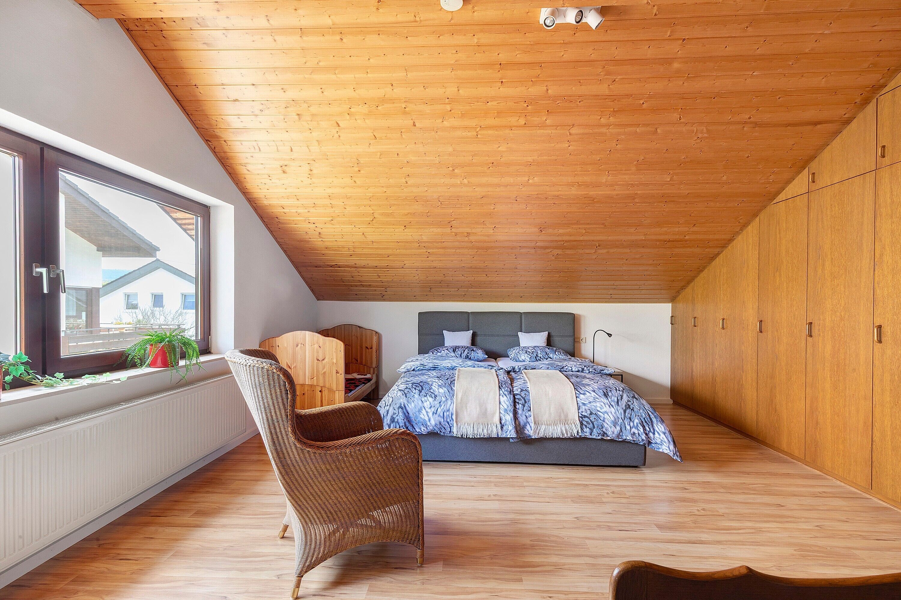 Un dormitorio con techo inclinado, una cama con colcha de rayas azules y blancas, una silla de mimbre, un armario de madera y una ventana con vistas a edificios.