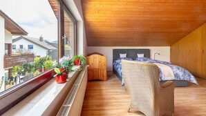 A bedroom with a large window, wooden ceiling, a bed with blue bedding, a wicker chair, and a small wooden cabinet. - Apartment Teckblick with Wi-Fi (Kirchheim unter Teck)