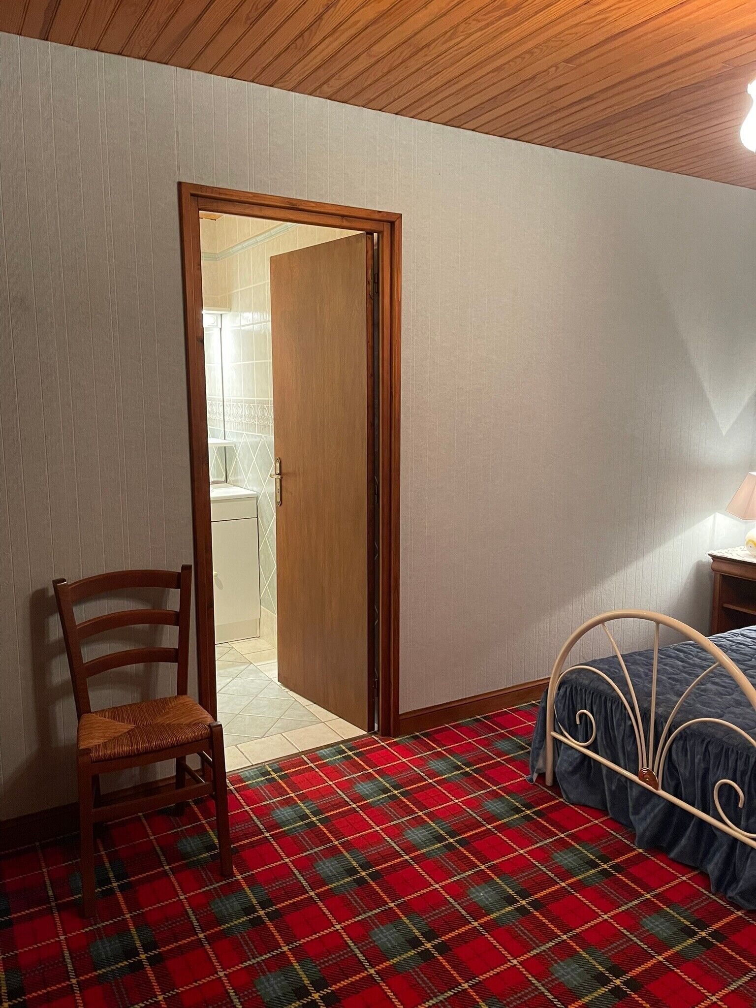 Habitació amb un rug plaidet, una porta de fusta, un fermall de llit metàl·lic i una cadira de fusta.