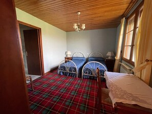 Une pièce avec deux lits en métal, un plafond en bois, un sol à carreaux rouges et verts, une nappe en dentelle blanche et une fenêtre avec des rideaux.