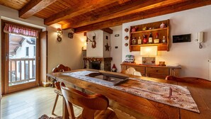 Een houten eettafel met stoelen, een houten plafond, een open haard en een plank met flessen.