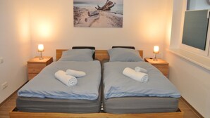 A double room with two beds, each with a pillow and a rolled towel. There are bedside tables with lamps and a painting of a beach scene on the wall. - Vacation apartment Meer-Zeit in Lubmin (Lubmin)