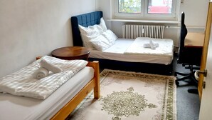 Quarto pequeno e arrumado com duas camas de solteiro, uma mesinha de cabeceira de madeira, uma escrivaninha com cadeira e um radiador.