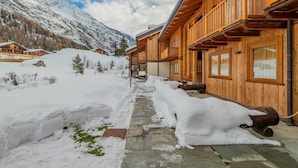 A wooden lodge with a stone pathway, snow-covered ground, and mountains in the background. - APT JARDIN DES ALPES - Let yourself be enchanted by the magic of the mountains (La Thuile)