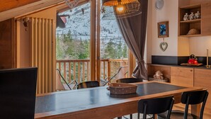 A wooden dining table with chairs, a view of trees through large windows, a hanging light fixture, and a kitchen area with cabinets and a kettle. - APT JARDIN DES ALPES - Let yourself be enchanted by the magic of the mountains (La Thuile)