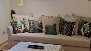 Una sala de estar con sofá, una mesa de centro, un tapete estampado, un letrero luminoso con forma de palmera y una ventana con vistas.
