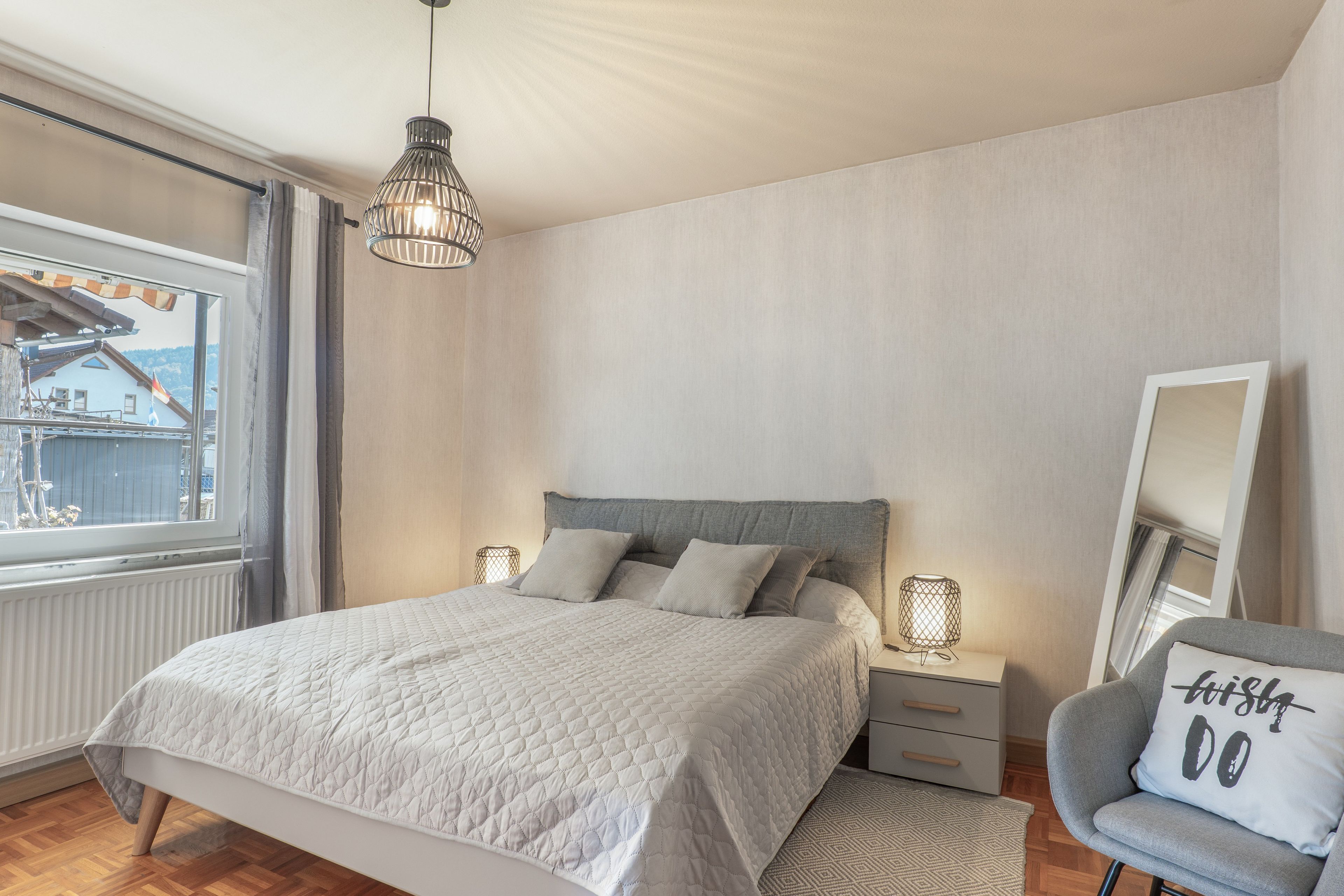 Un dormitorio con cama, lámparas de noche, una silla, un ventanal y vista a un edificio.