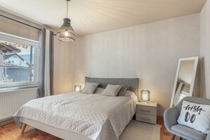 Ein Schlafzimmer mit einem Bett, Nachttischlampen, einem Sessel, einem Fenster und Blick auf ein Gebäude.