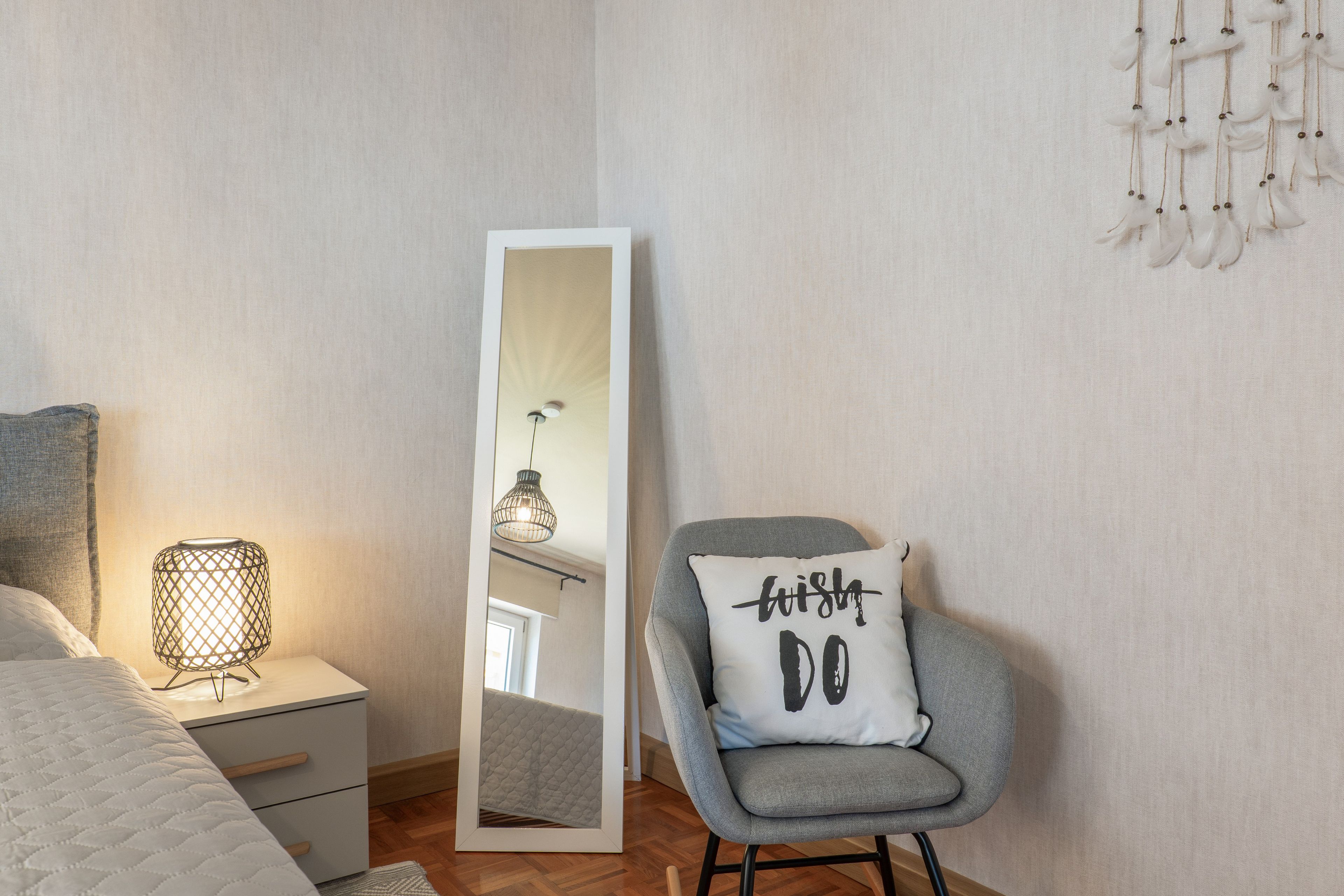 A bedroom with a bed, a nightstand, a lamp, a chair with a pillow, a mirror, and a wall with a dreamcatcher.