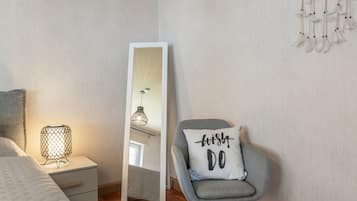 A bedroom with a bed, a nightstand, a lamp, a chair with a pillow, a mirror, and a wall with a dreamcatcher.