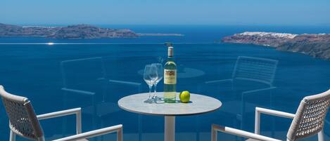A glass table with a bottle and a glass, two wicker chairs, and a view of the sea and cliffs.