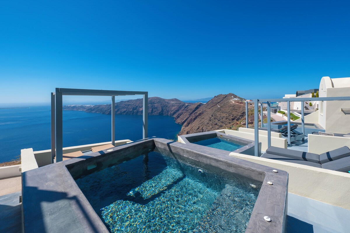 A rooftop infinity pool with clear blue water, white architecture, and a view of the sea and mountains.
