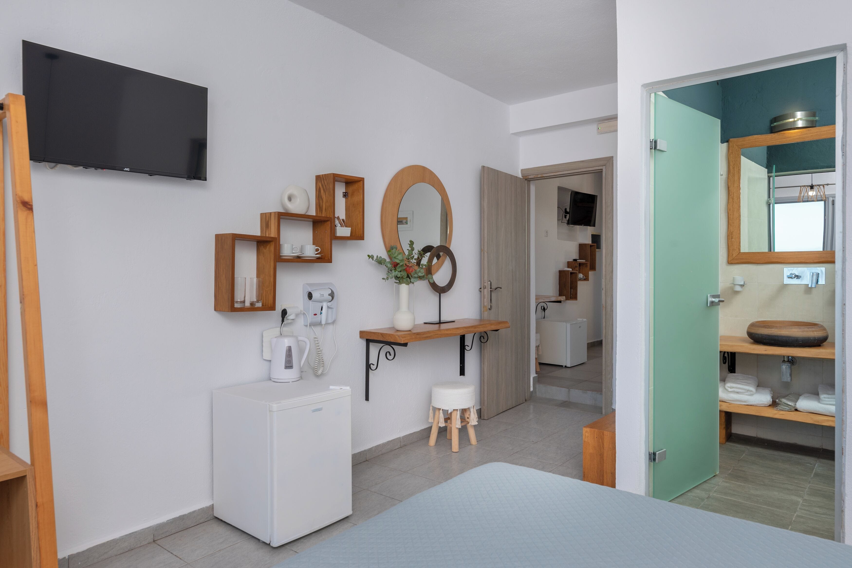 A modern hotel room with a flat-screen TV, a compact refrigerator, a wooden vanity with a mirror, and a small stool.