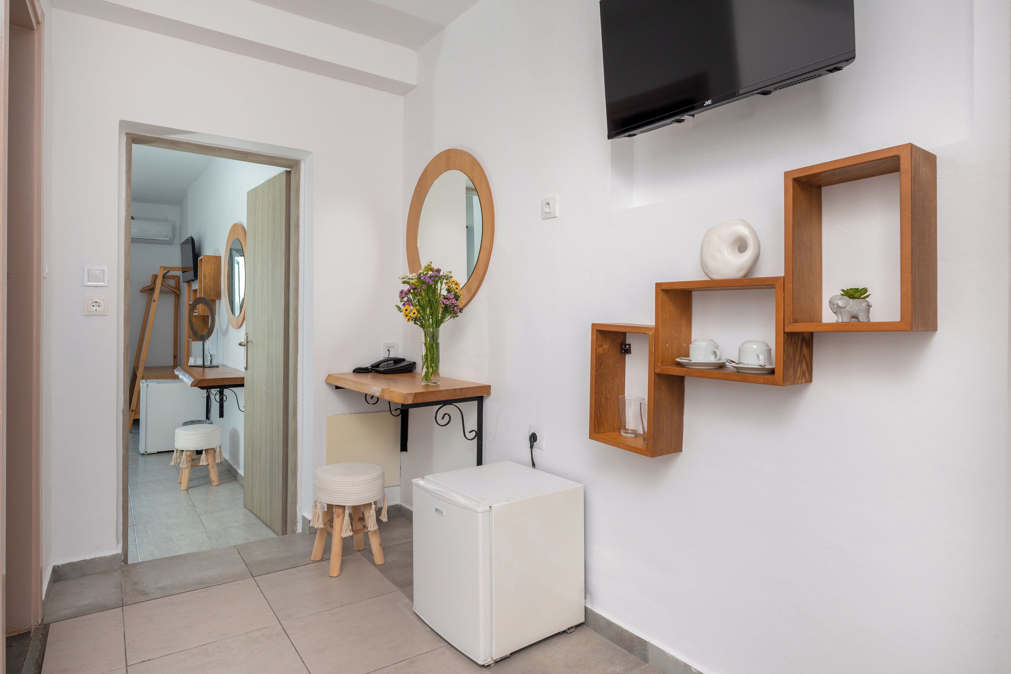 Een moderne kamer met een flatscreen televisie, houten planken, een kleine koelkast, een spiegel en een bureau met een bloevas.
