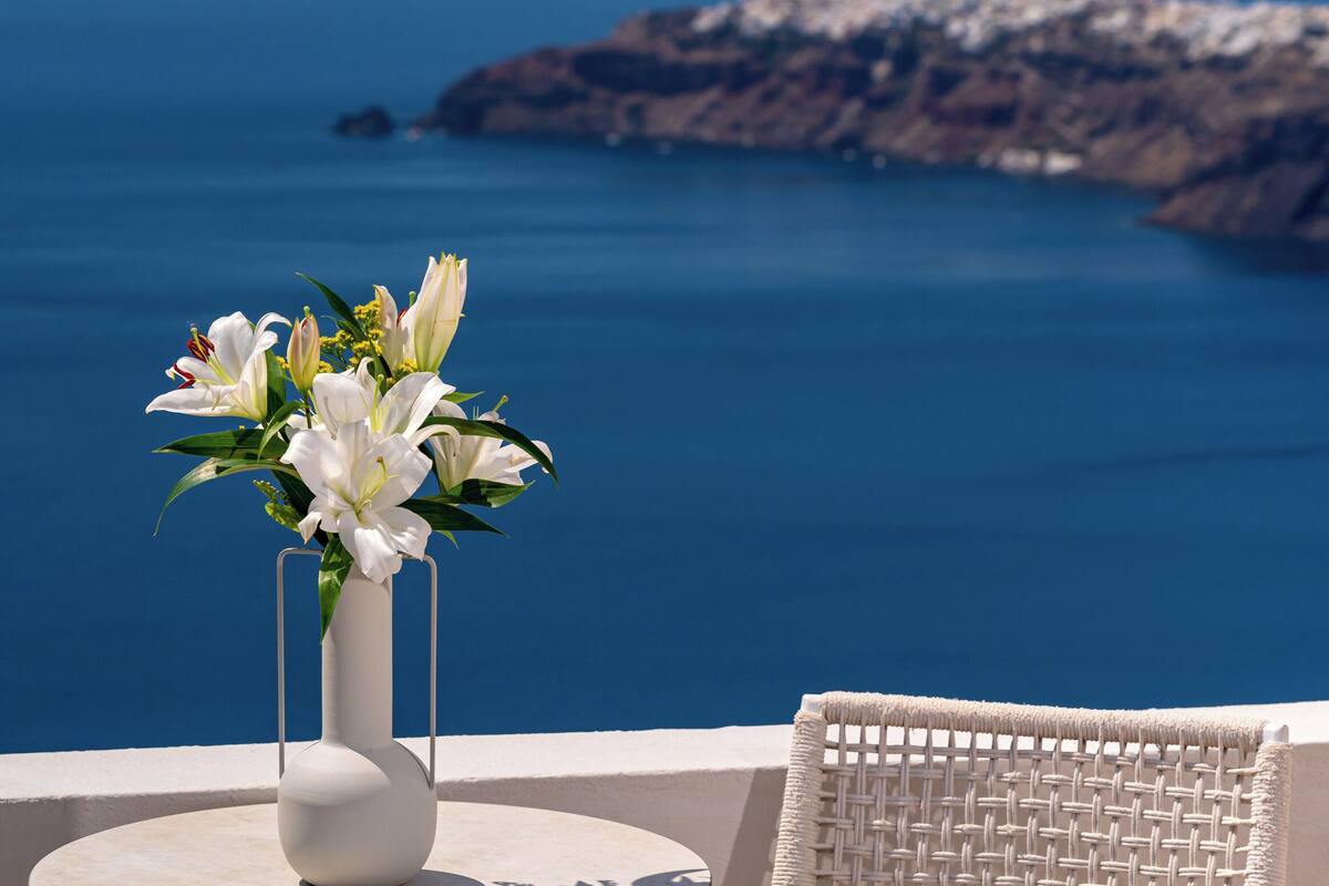 A round table with a white vase of flowers, a wicker chair, and a view of the sea and rocky coastline.