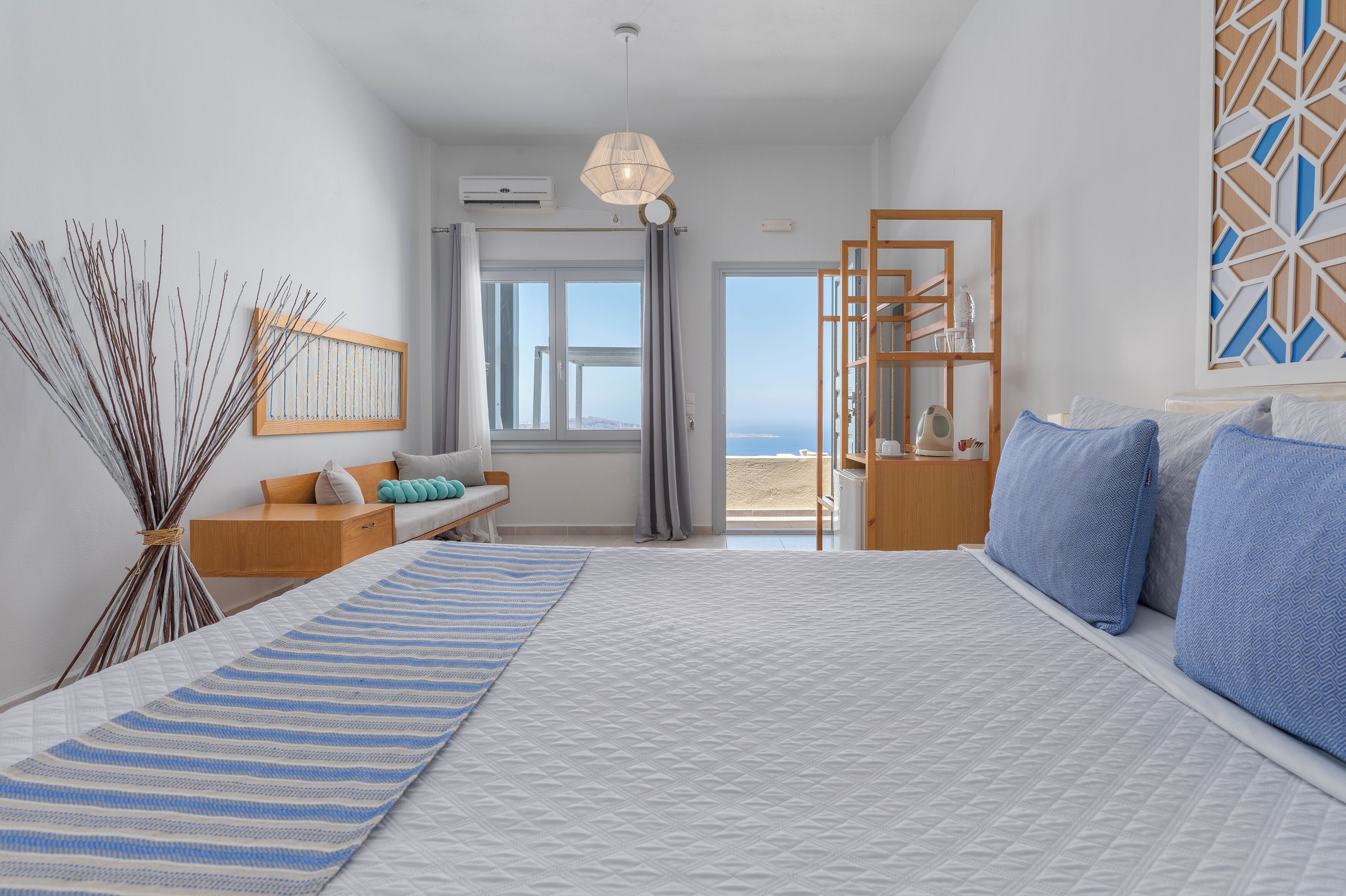 A bedroom with a large bed, a bedside table, a nightstand, a window with curtains, a view of the sea, and a decorative wall panel.