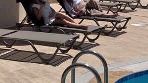 A poolside area with lounge chairs and umbrellas, a stone building with a balcony, and a palm tree in the background.