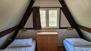 Um quarto de sótão pequeno e triangular com duas camas de solteiro, um armário de madeira e uma janela com cortinas.
