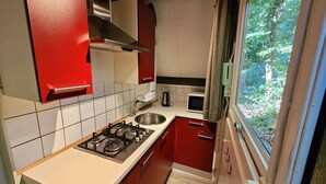 Cozinha compacta com fogão a gás, pia e micro-ondas. Armários vermelhos e uma janela com vista para o verde são visíveis.